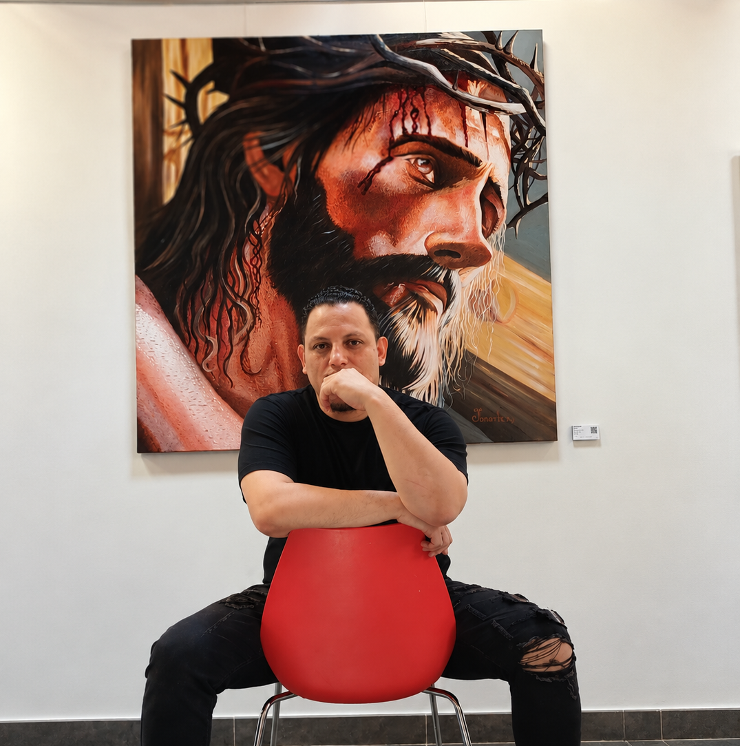 Jonarte Rodríguez sentado frente a su obra “Mesías” en la sala de exposición