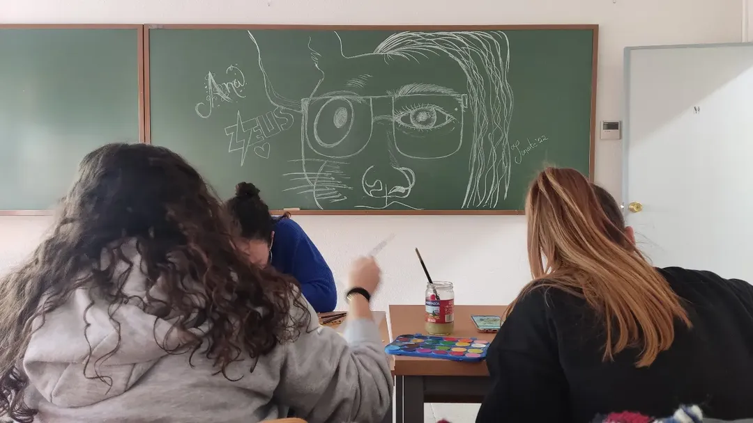 Alumnas dibujando en un aula frente a un retrato dibujado a tiza en la pizarra por JonArte